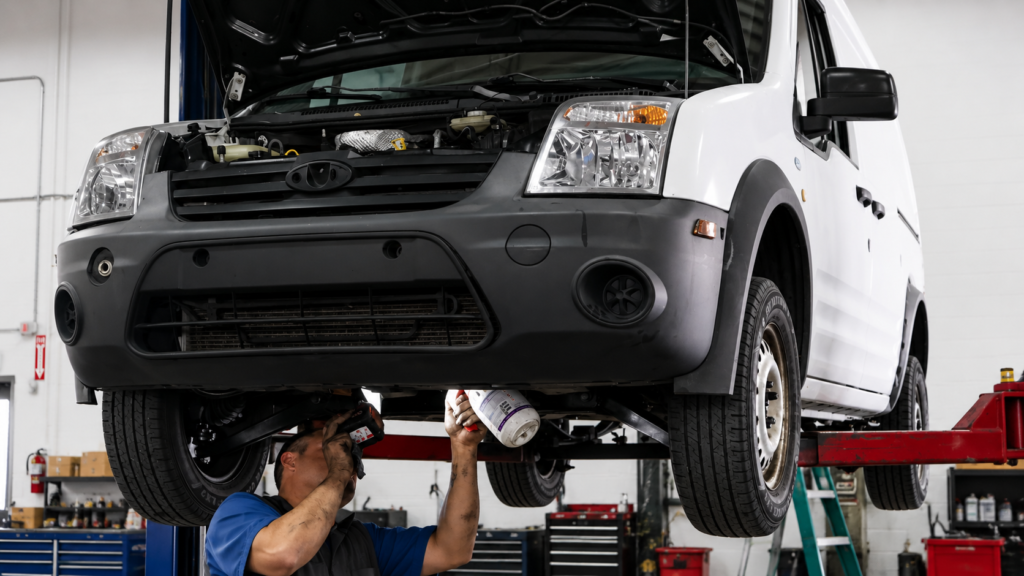 Technician performing kingpin and front-end repair service on a white commercial work van lifted in an automotive repair shop, inspecting and maintaining suspension and steering components for fleet vehicle safety and performance.