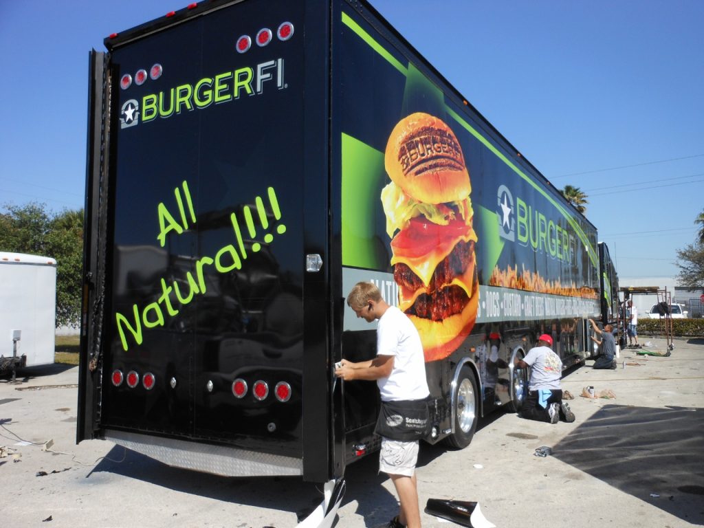 Technicians installing custom vehicle wrap on BurgerFi trailer with large food graphics